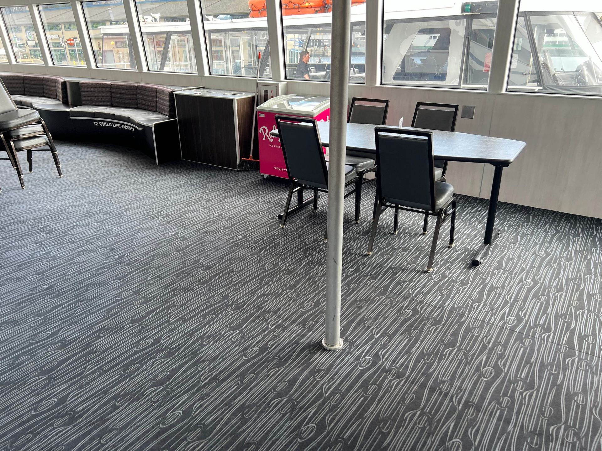 Interior of a boat with gray carpet, seating, tables and windows.