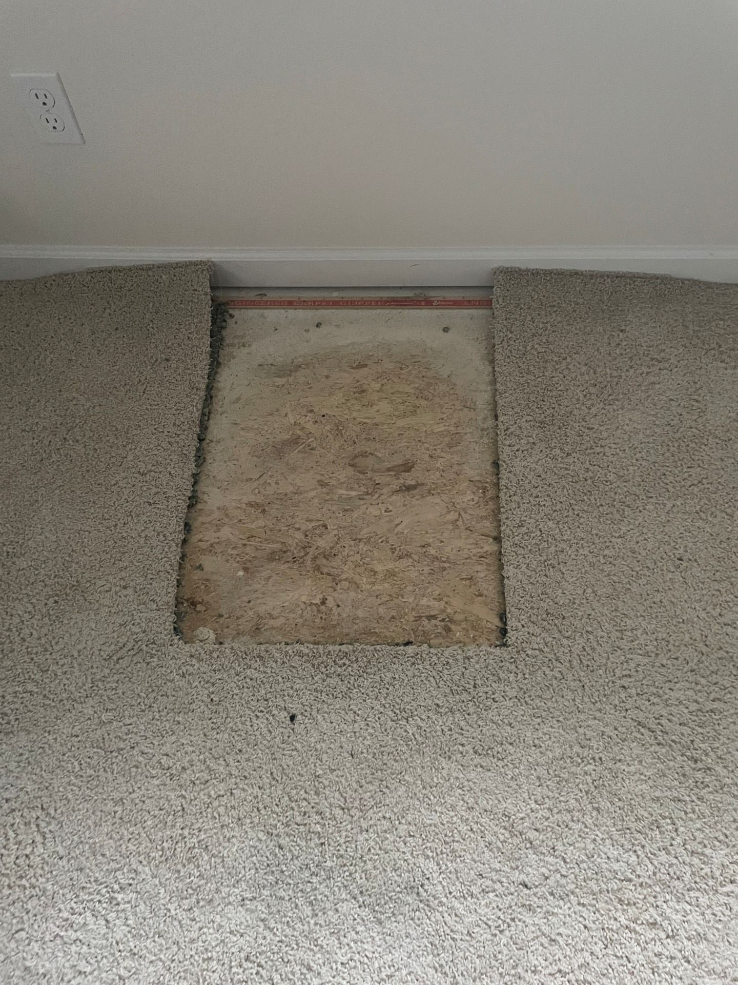 Hole cut out of light-colored carpet, revealing the subfloor. Edges of carpet are exposed along the top.