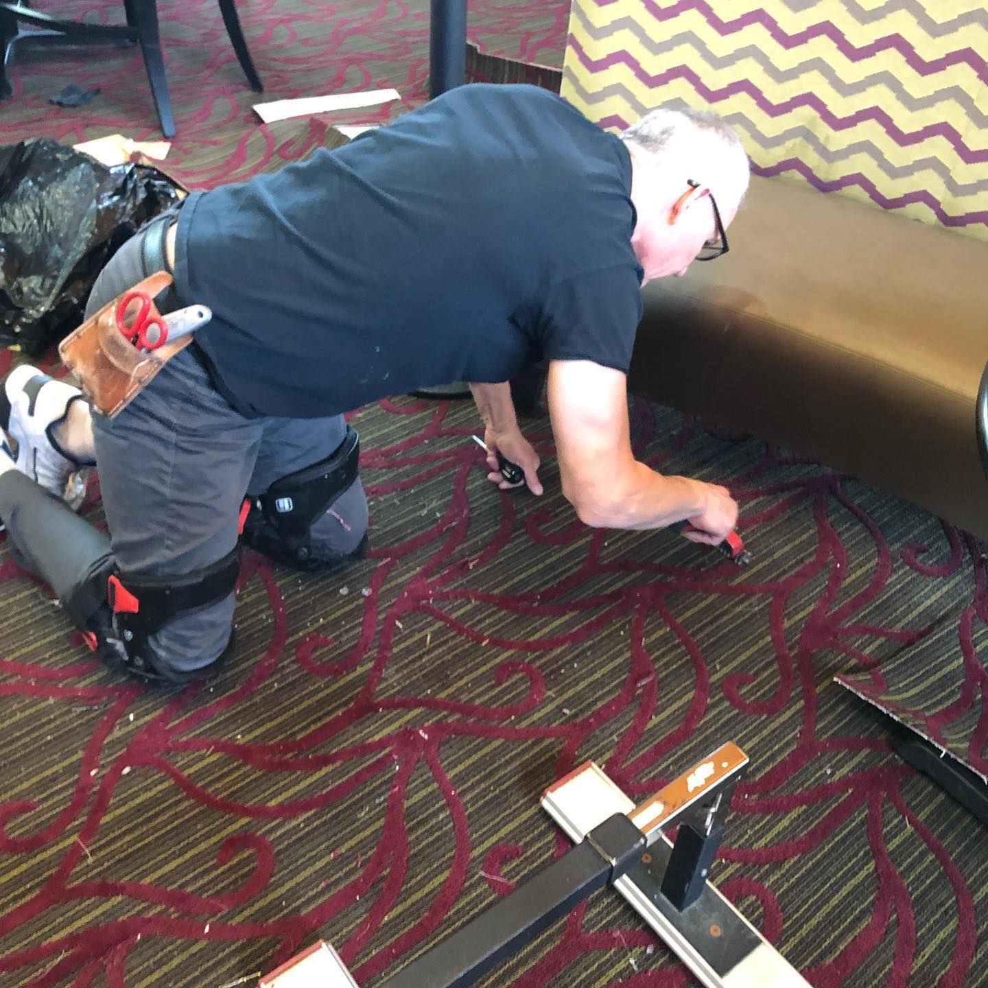 Man on knees cutting carpet with a tool. He wears kneepads, a tool belt, and is in a room with patterned carpet.