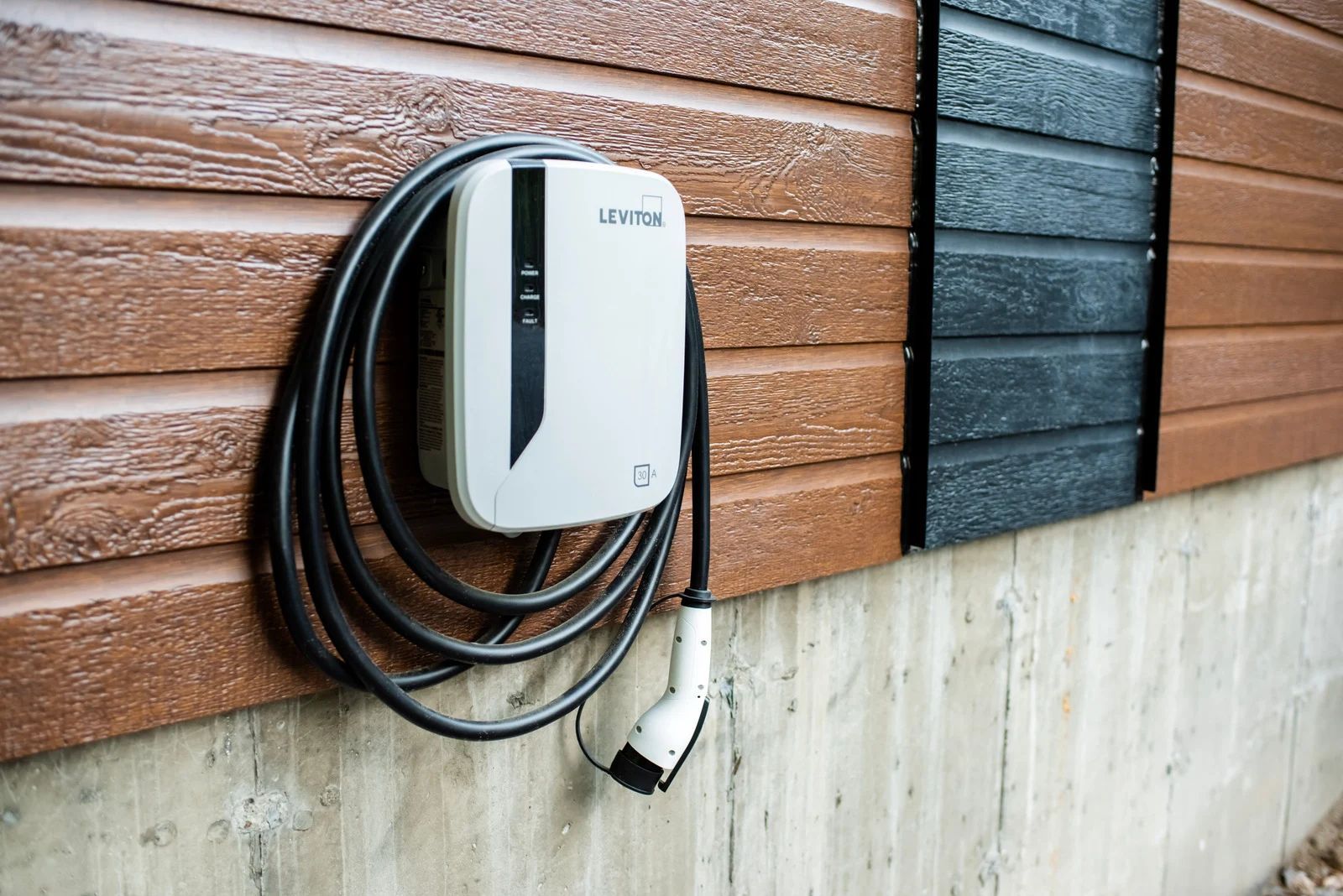 White EV charger mounted on a brown and black siding wall, with a coiled black cable and charging plug.