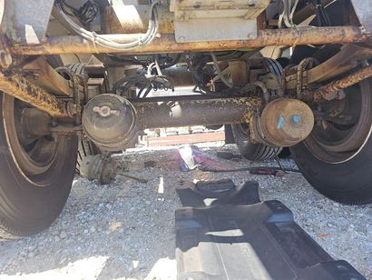 Undercarriage of a rusty truck showing axles, wheels, and brake components on a gravel surface.
