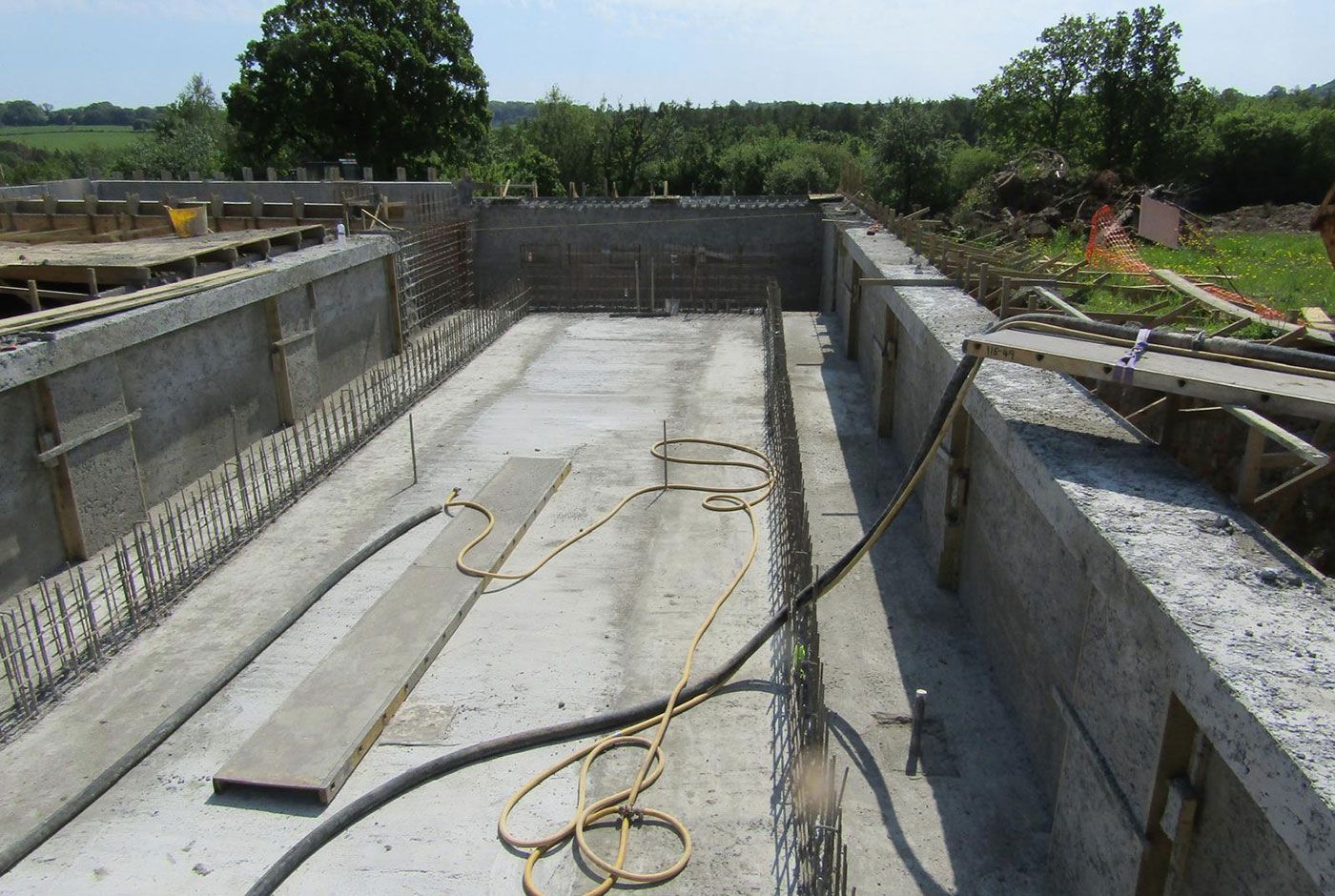 Open air concrete foundation under construction with various hoses on the floor, trees in the background.