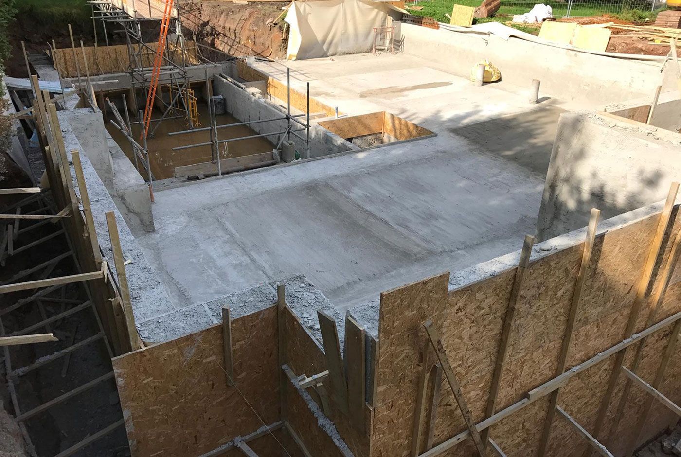 Construction site on a sunny day with a concrete foundation surrounded by wooden formwork and scaffolding.