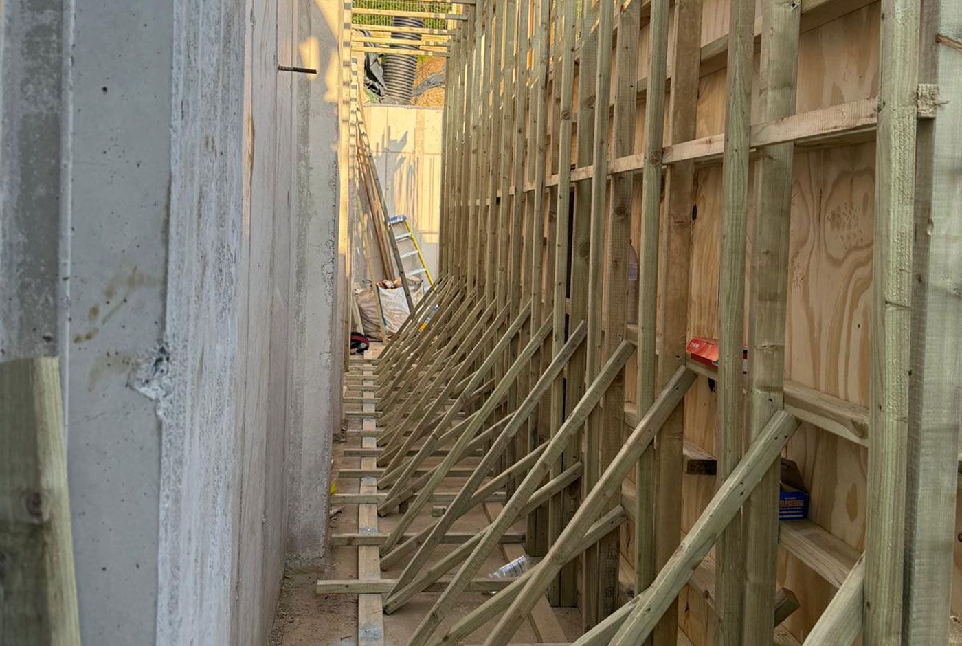 Narrow construction walkway between concrete wall and wooden framing, with braces and scaffolding.