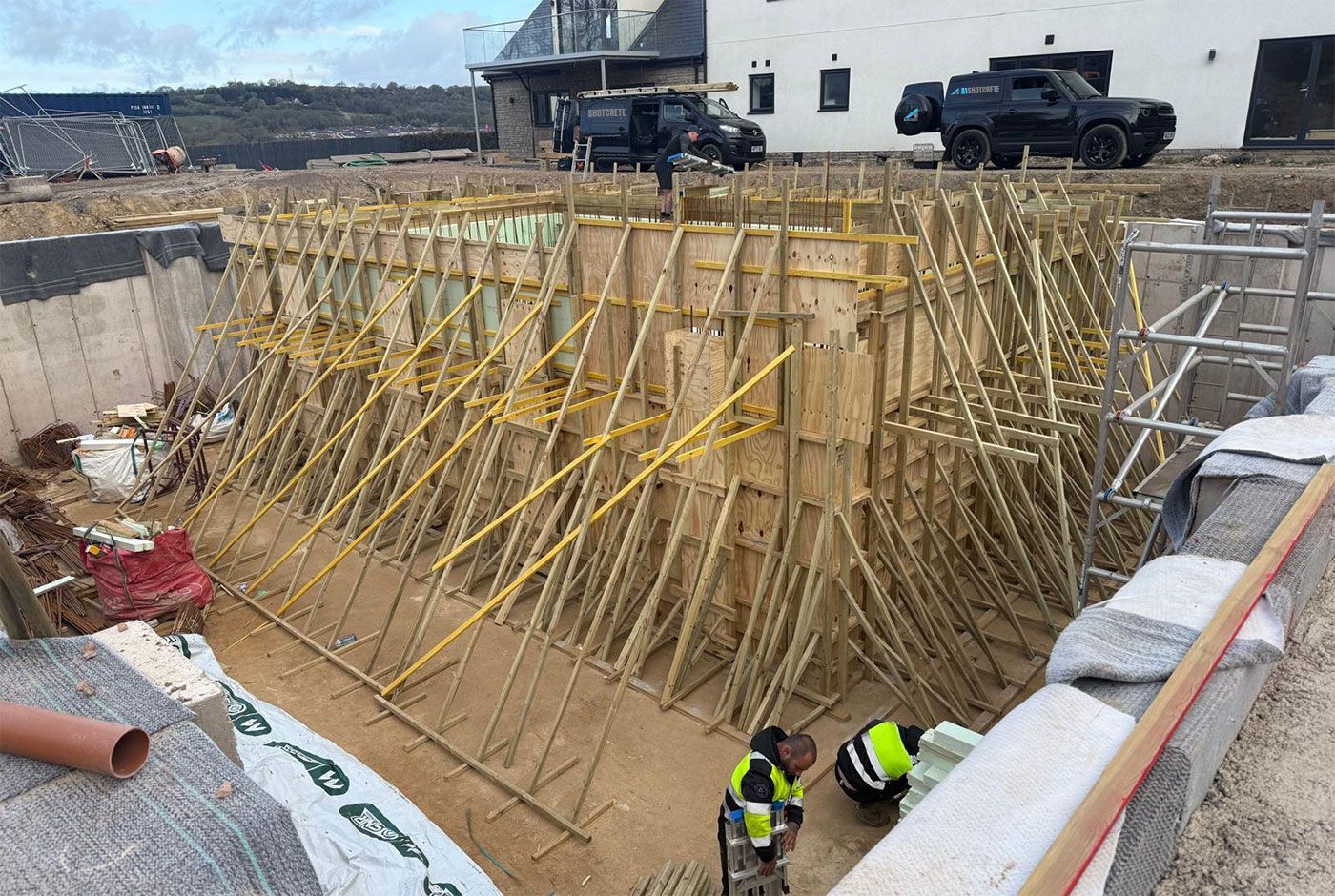 Construction crew working in a deep excavation site with braced wooden formwork, vehicles and a house in the background.