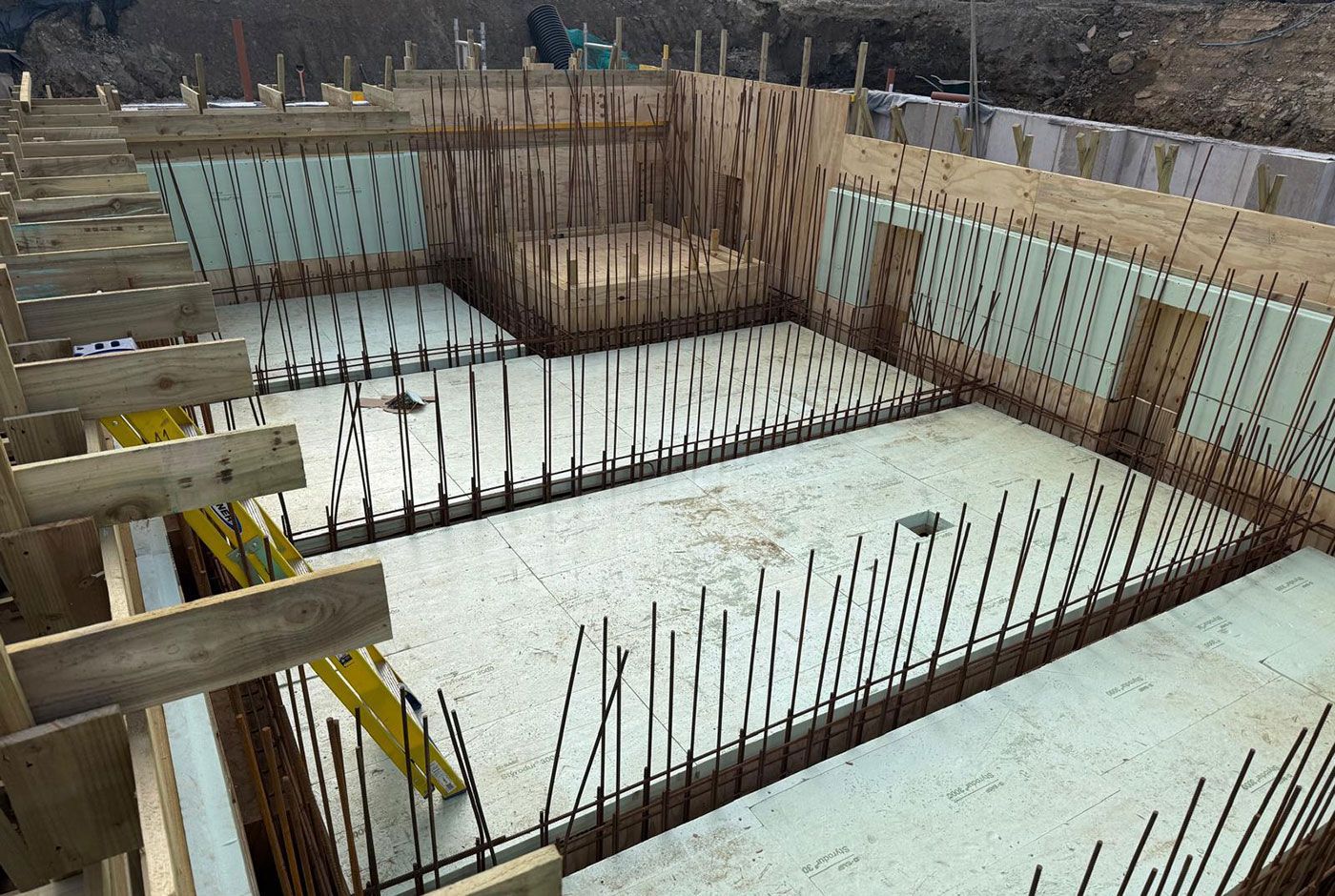 Concrete foundation pit with steel rebar and formwork at a construction site.