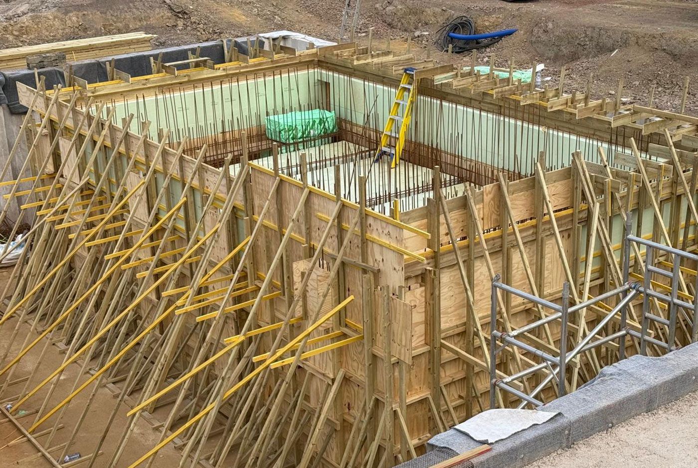 Foundation excavation with wooden formwork and yellow bracing and scaffolding on a construction site.