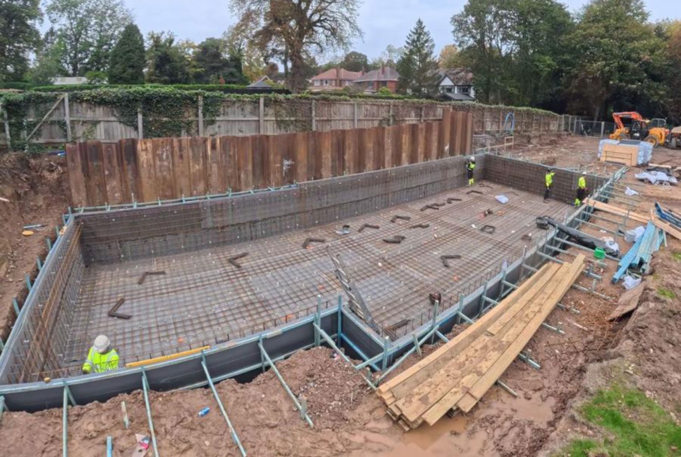Construction site in a residential garden with a large reinforced concrete pool shell and workers in green PPE.