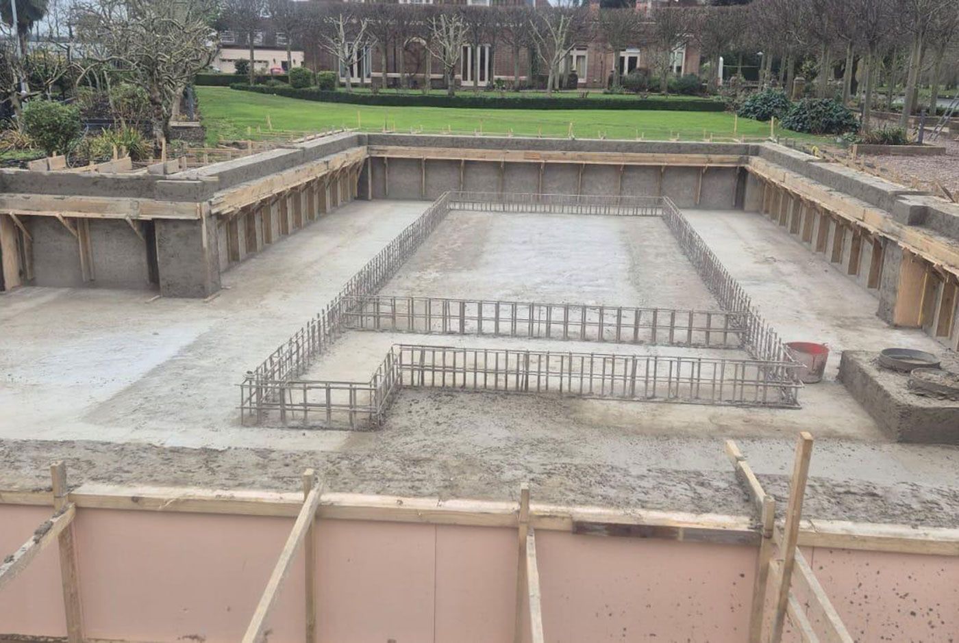 Excavated concrete foundation pit with rebar frames, surrounded by retaining walls, property and trees in the background.