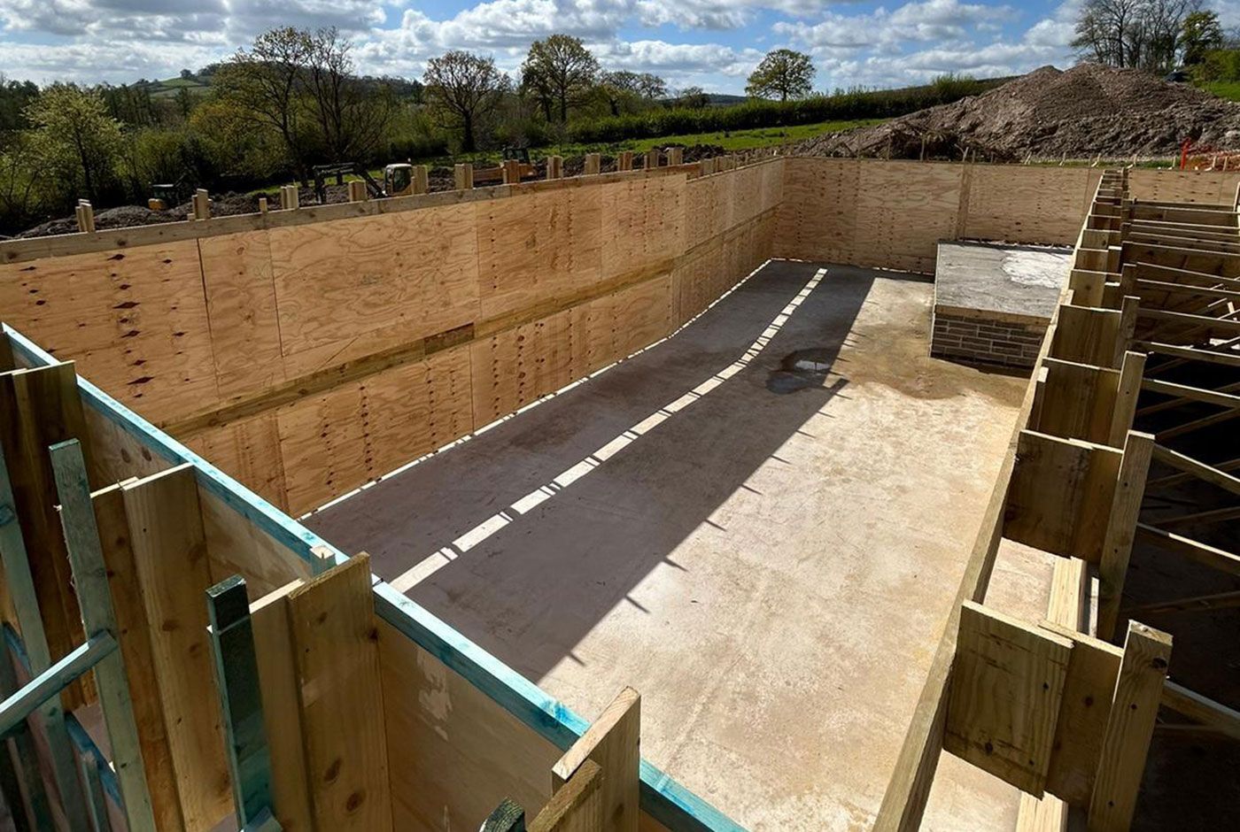 Construction site with a deep excavation, timber retaining walls, and countryside in the background.