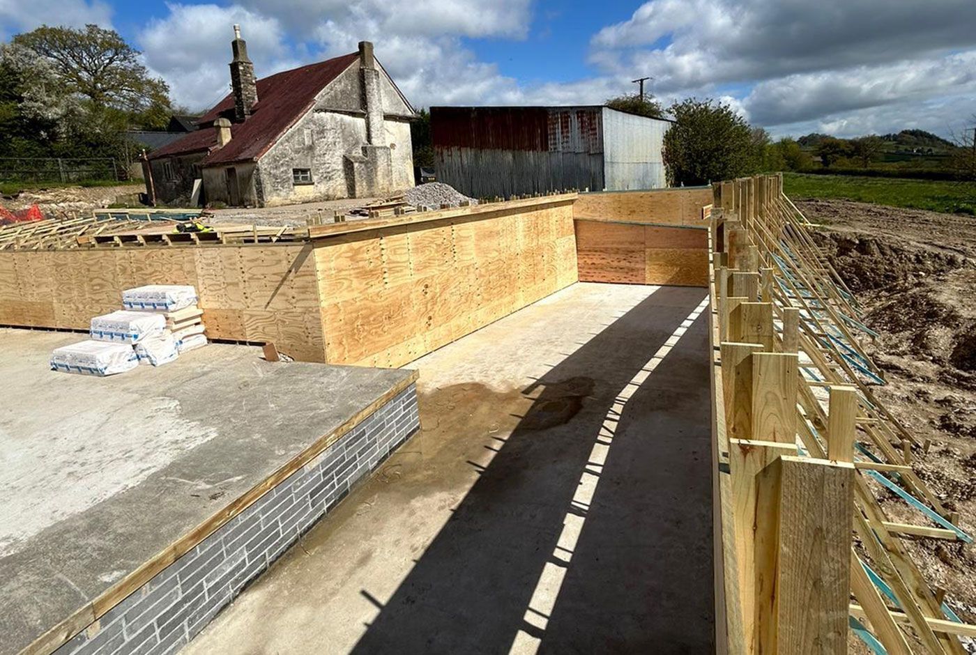 Construction site with timber foundation walls and bracing, a house and barn in the background, fields beyond.
