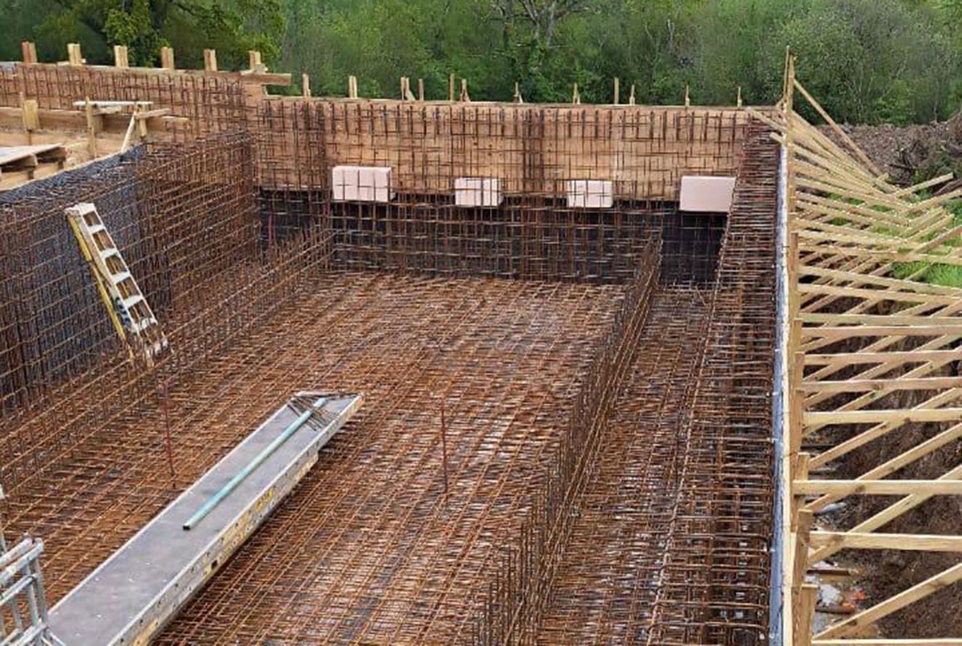 Partially built foundation structure with timber scaffolding and rebar grid, leafy trees in the background.