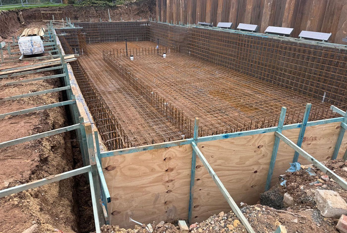 Construction site with a large rectangular foundation pit  with rebar, reinforced by wooden formwork.