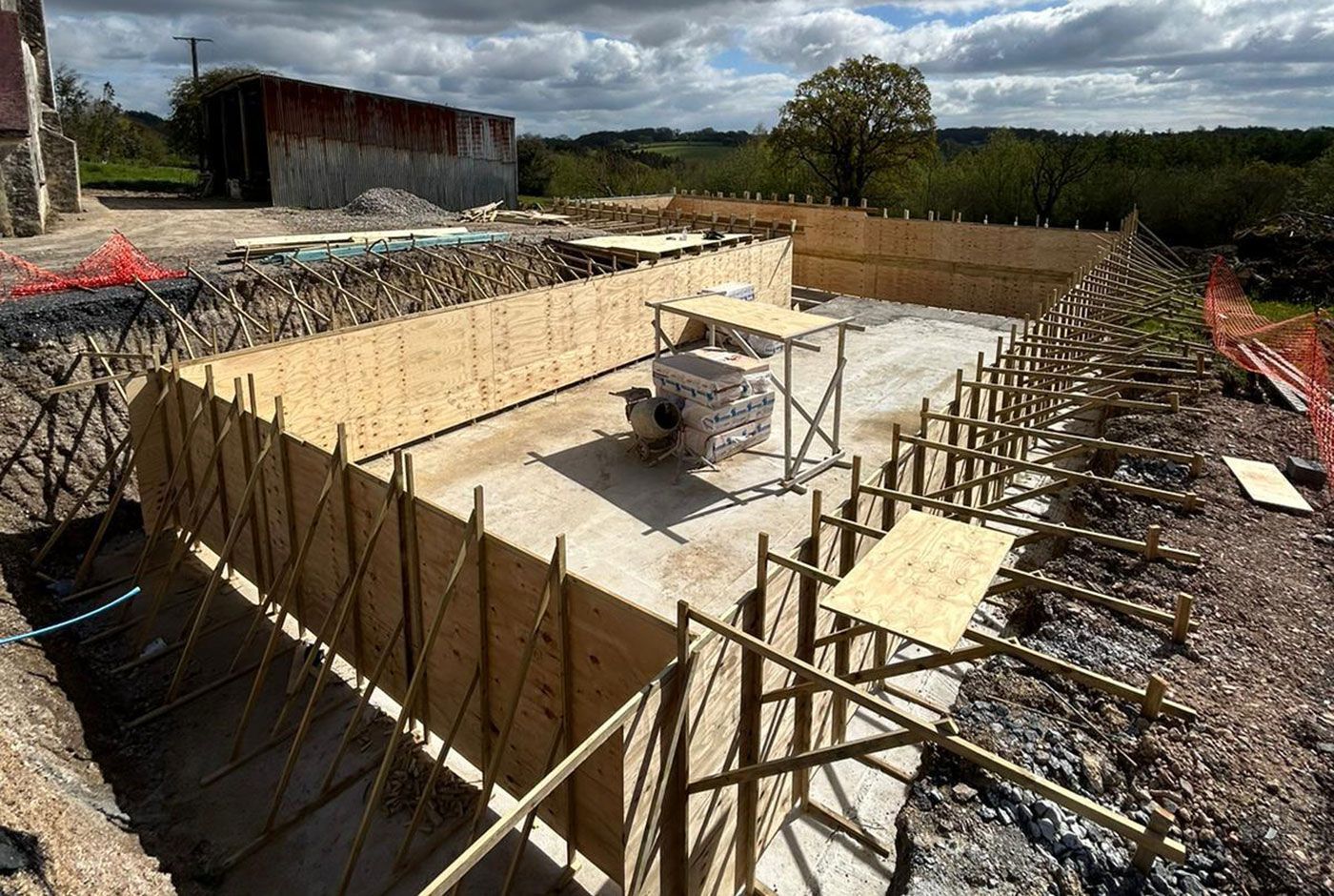 Construction site with a large foundation pit, concrete base, wooden formwork, a barn, countryside and cloudy sky.