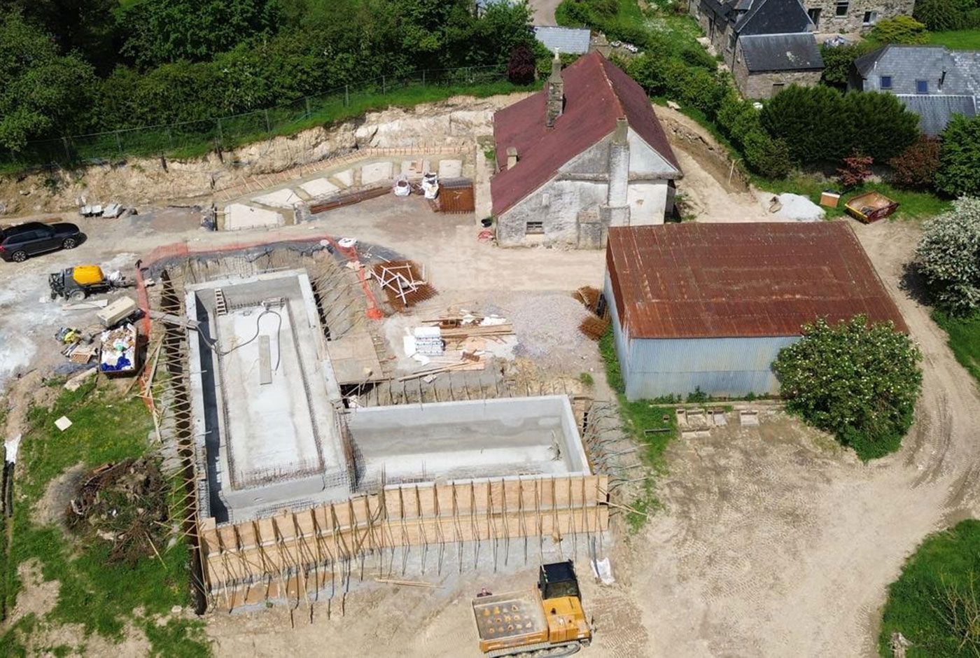 Aerial view of a construction site with concrete foundations, two red-roofed buildings, equipment and vehicles..