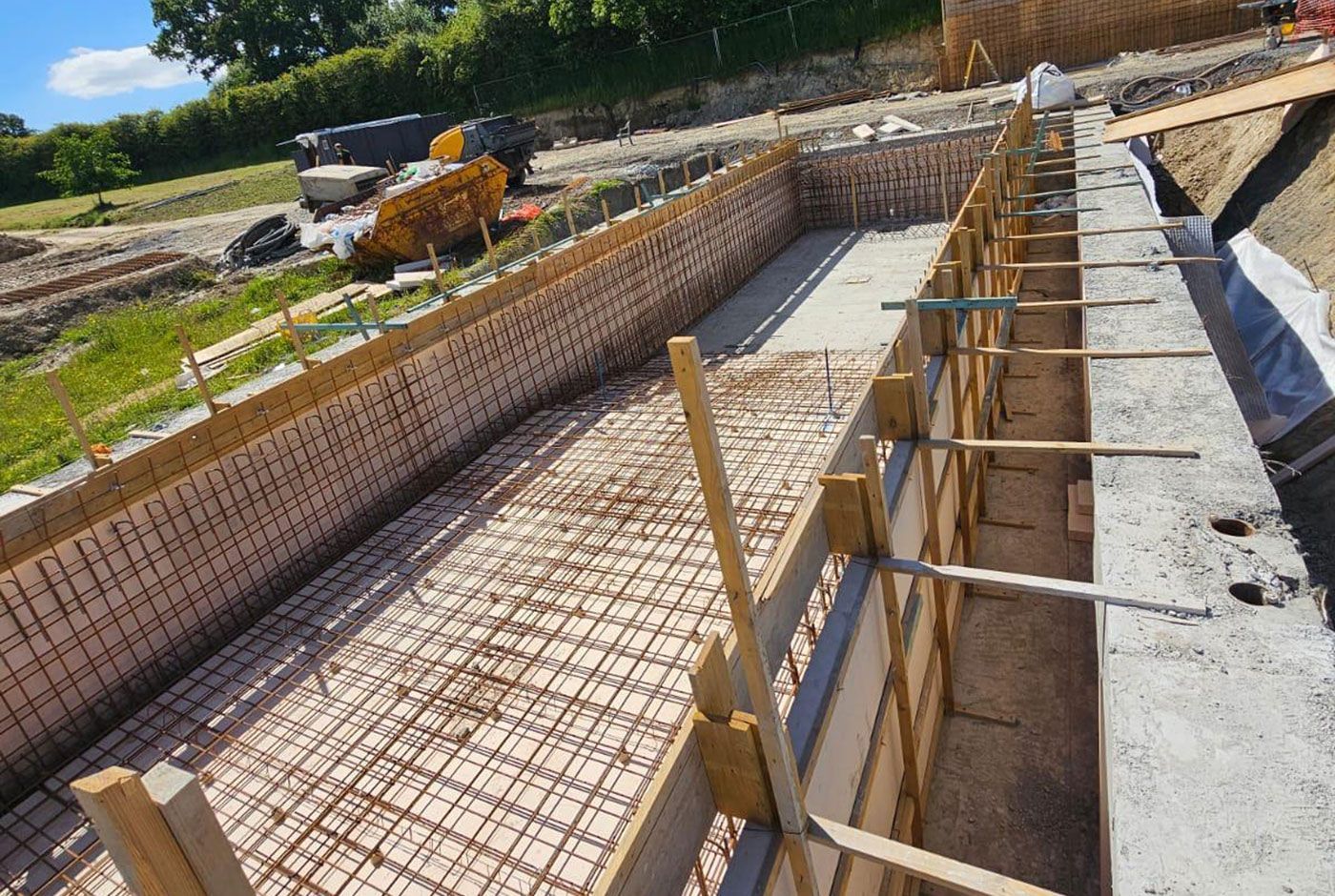 Concrete construction site with a rectangular foundation, rebar grid, and wooden formwork under bright sun.