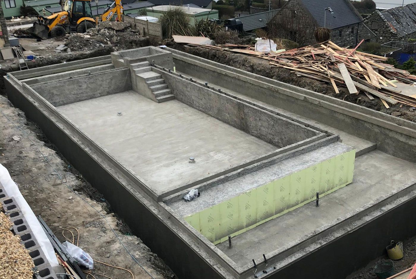 Concrete foundation of a swimming pool under construction, surrounded by excavation equipment and stack of lumber.
