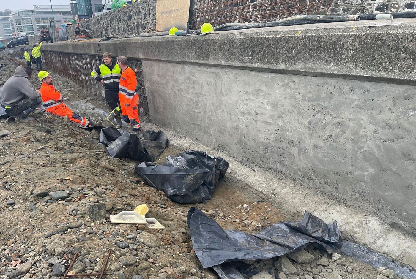 Workers in hi-viz safety gear taking a break on rough ground next to a concrete retaining wall.