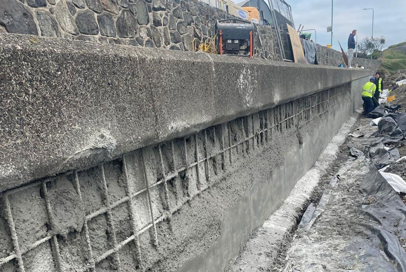 Construction workers at a retaining wall under repair with concrete over a mesh frame.