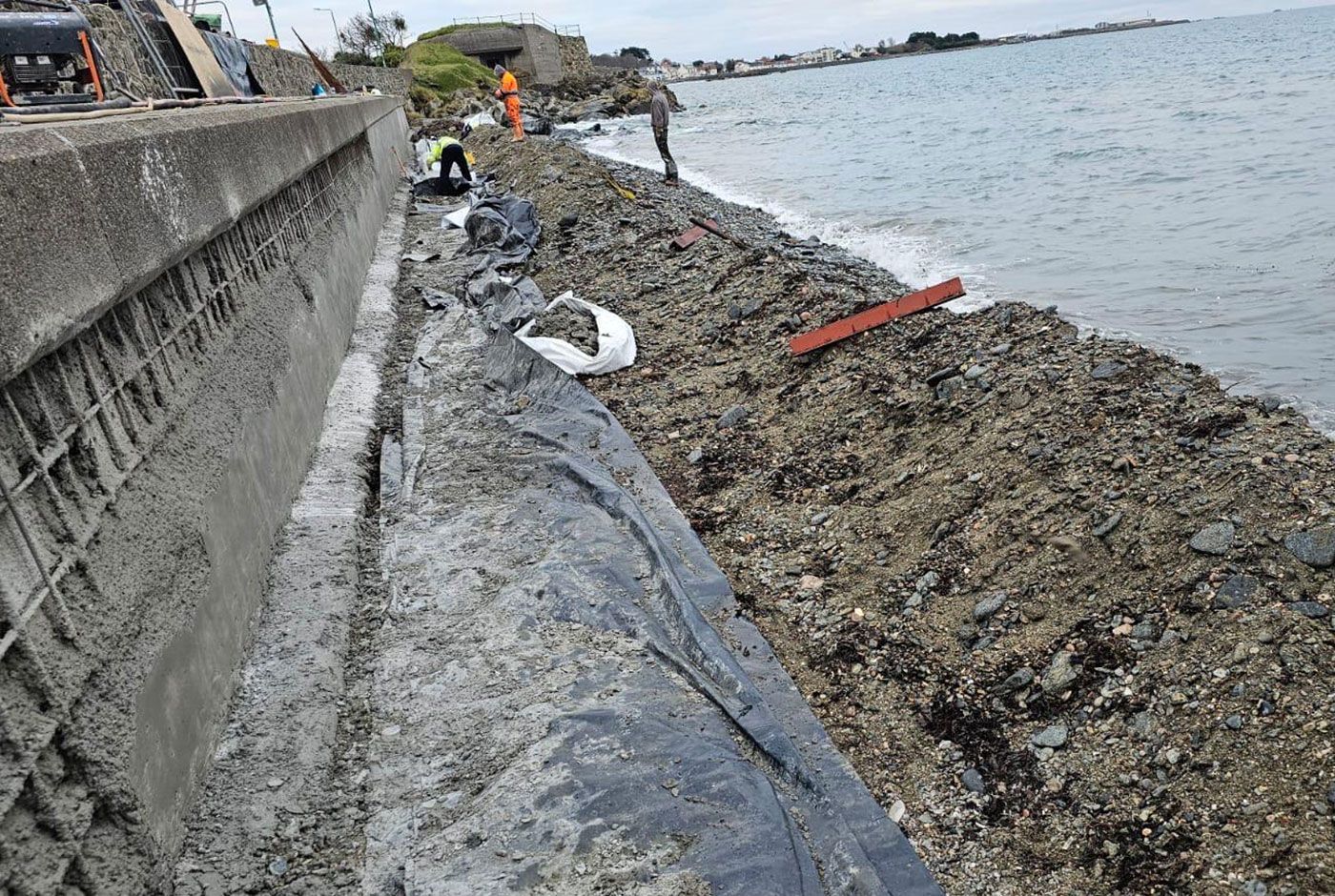 Rocky shoreline beside a concrete seawall that is being repaired with fresh concrete,  black tarpaulin along the waterline.