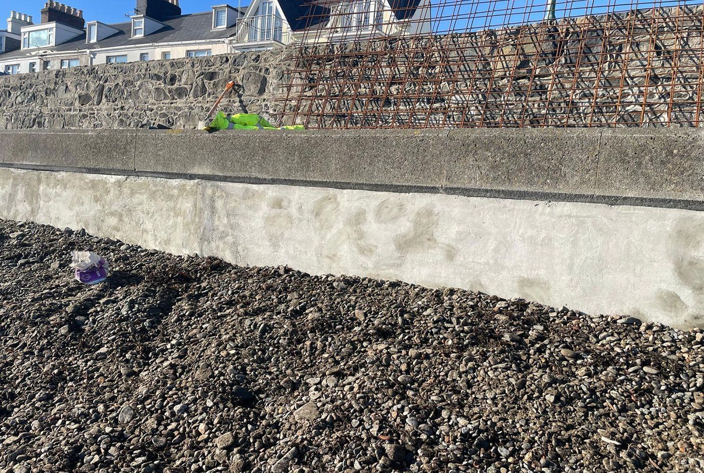 Coastal stone wall with cement base, mesh above, pebble beach below.