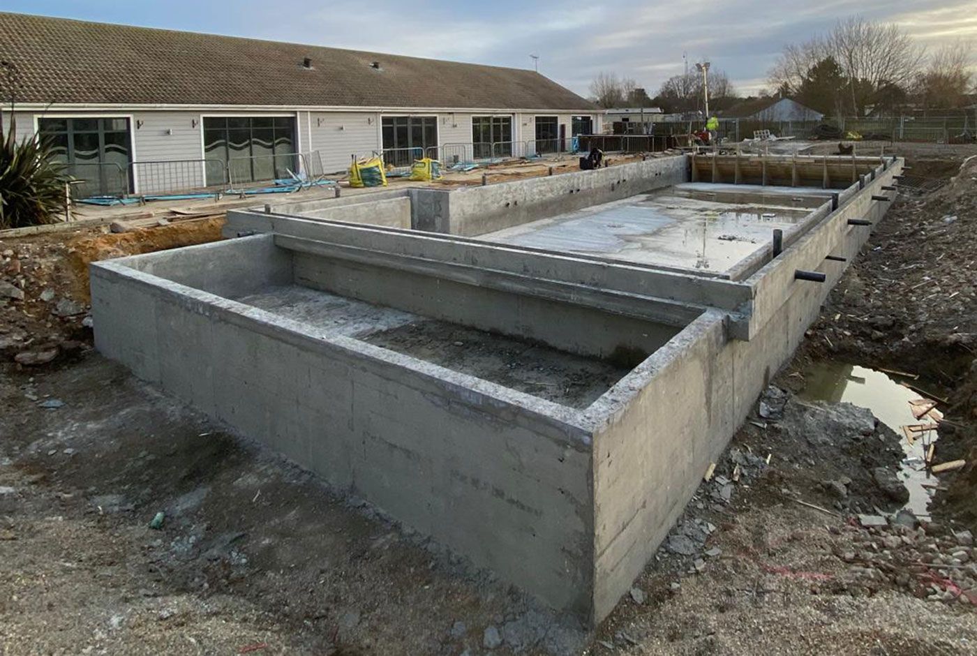 Concrete shell of a deck-level pool under construction in a muddy garden, with a single-storey building in the background.