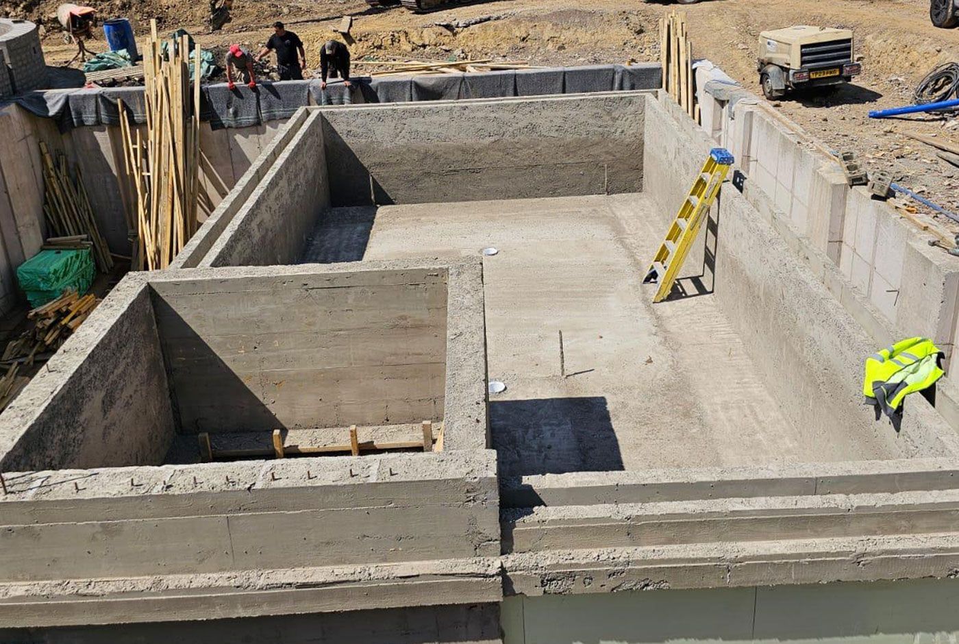 An outdoors deck-level pool shell under construction, with a ladder, materials and workers visible.