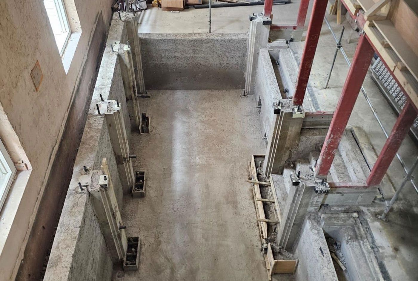 Overhead view of the shell of an indoor deck-level pool under construction, red steel supports and scaffolding.