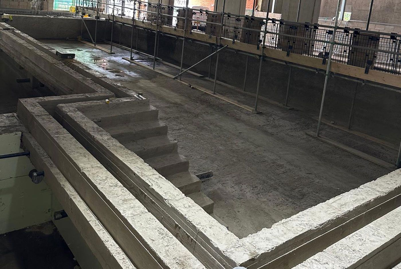 An indoor deck-level pool shell, almost finished with concrete walls, floor and steps, scaffolding and metal railings.