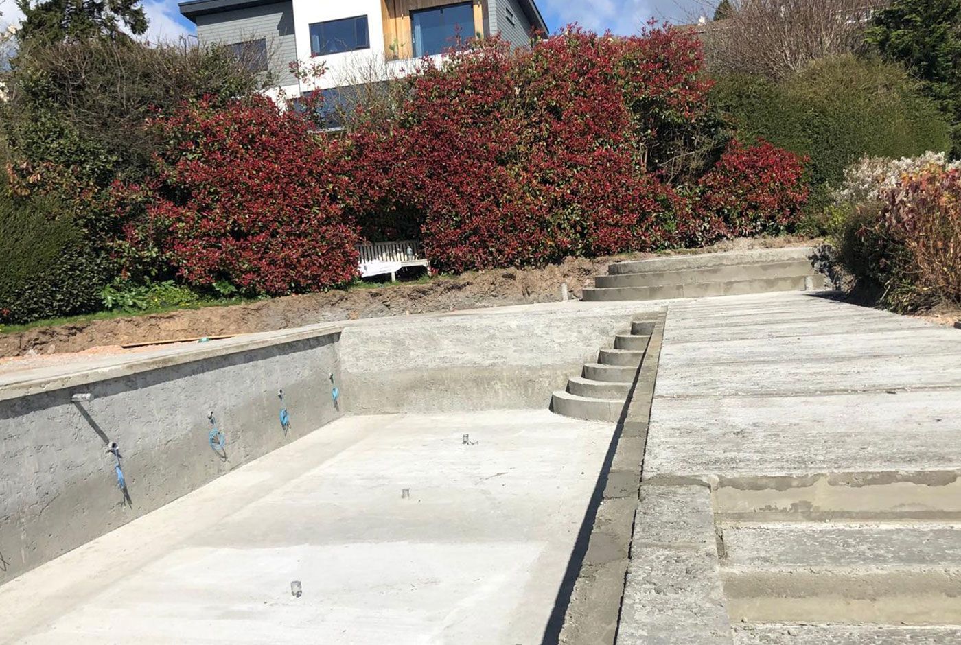 A completed deck-level swimming pool shell with steps down, mature shrubbery and a house in the background. 