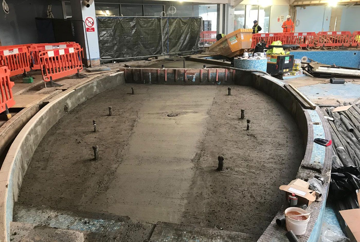 Large freeform pool under construction with concrete base, with workers, orange safety barriers and tools in an indoor site.