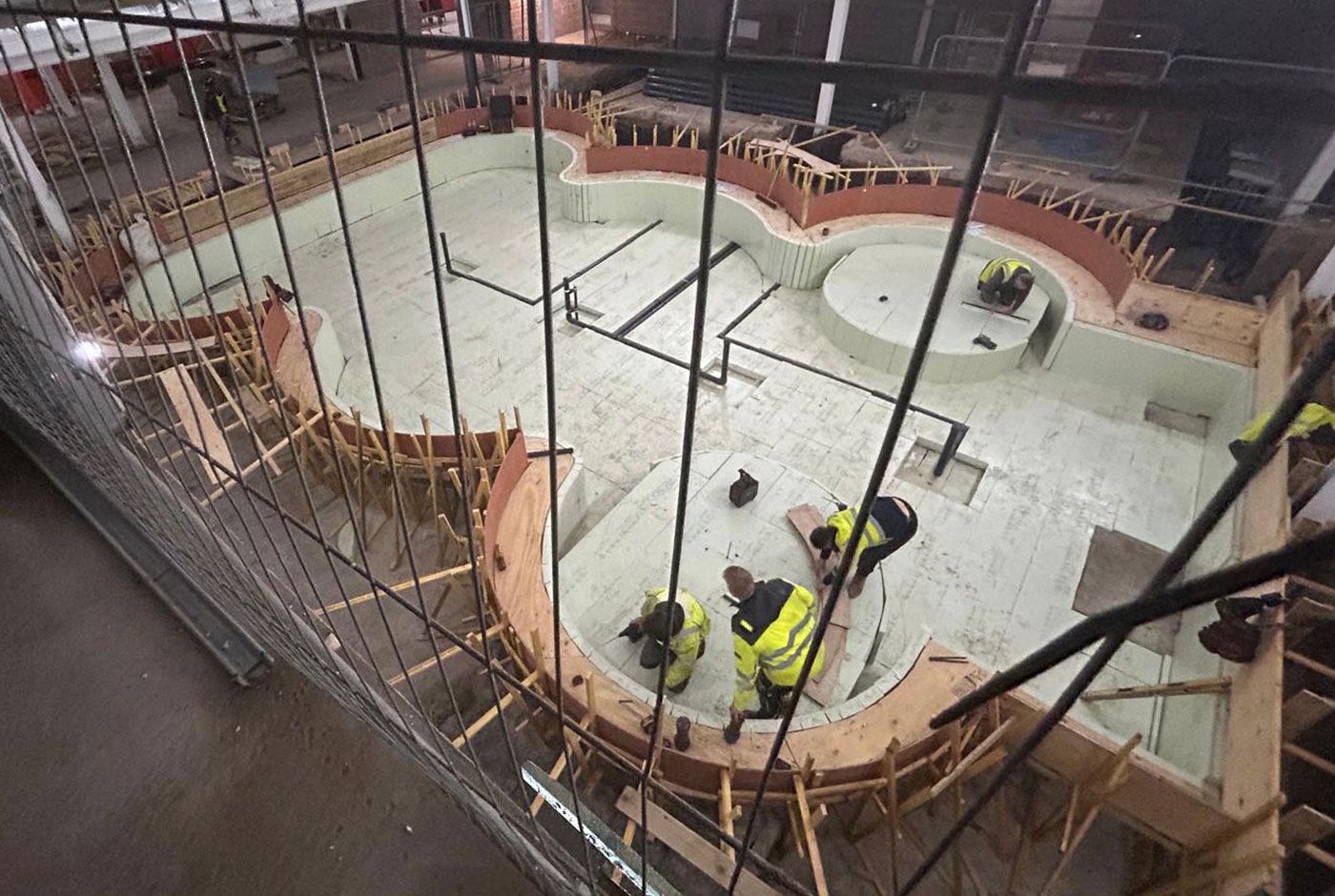 Overhead view of workers in hi-viz working on the site of a large indoor curved concrete pool under construction.