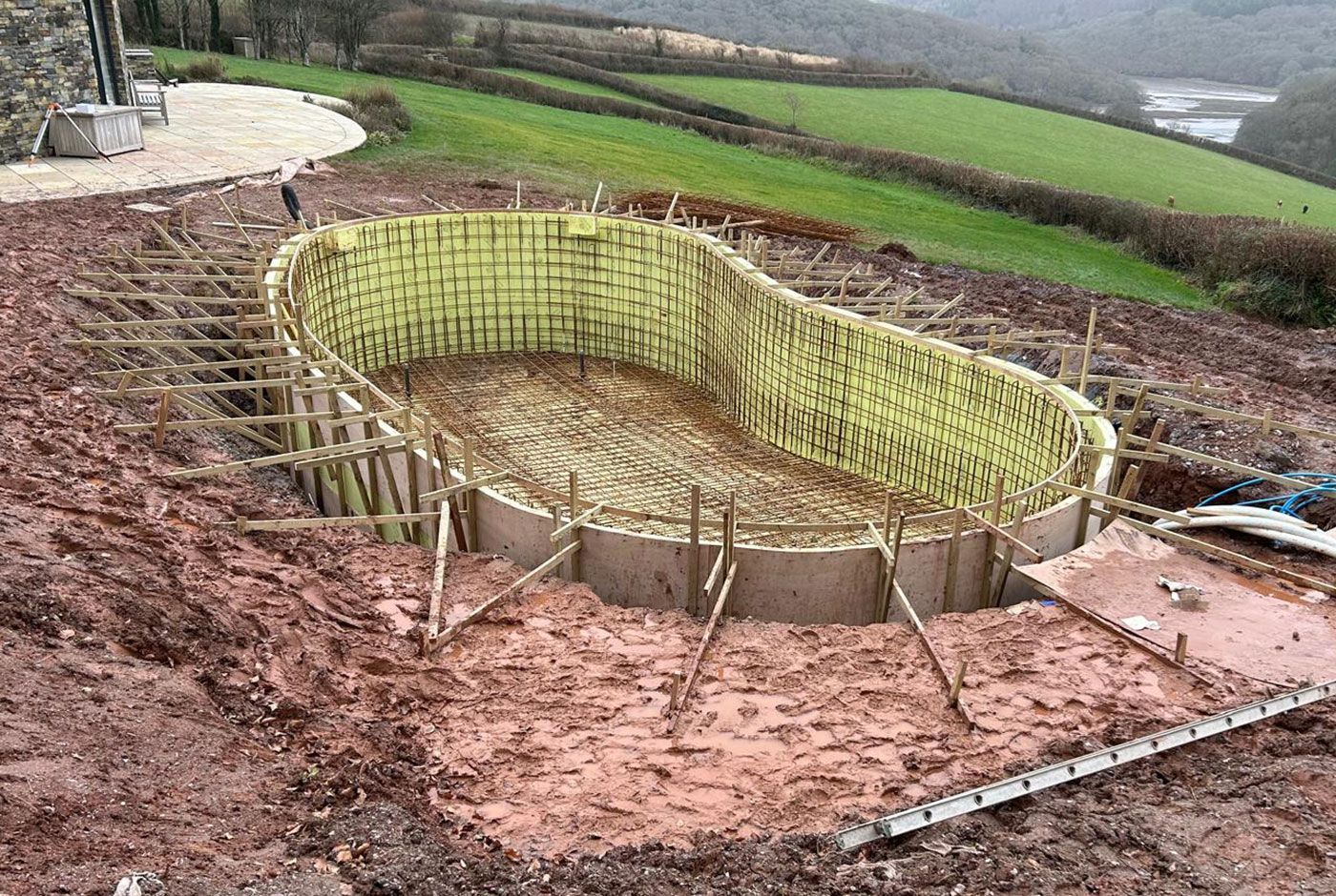The curved shape of a swimming pool under construction on a muddy location with green fields in the background.