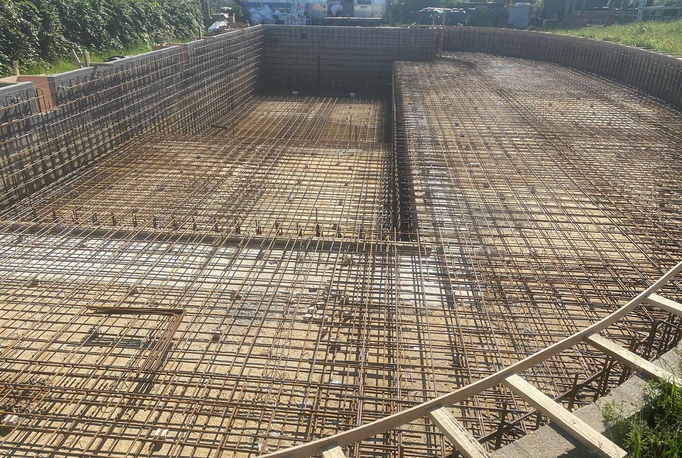 Large outdoor curved pool under construction with rebar grid and multi-level floor, surrounded by retaining walls.
