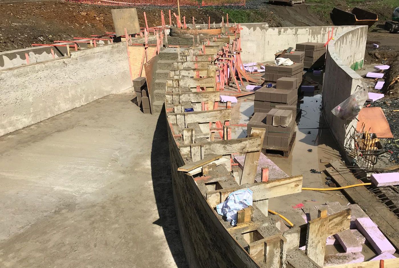 Construction site of a freeform pool with concrete walls and steps, wood formwork and stacked concrete blocks beside it.