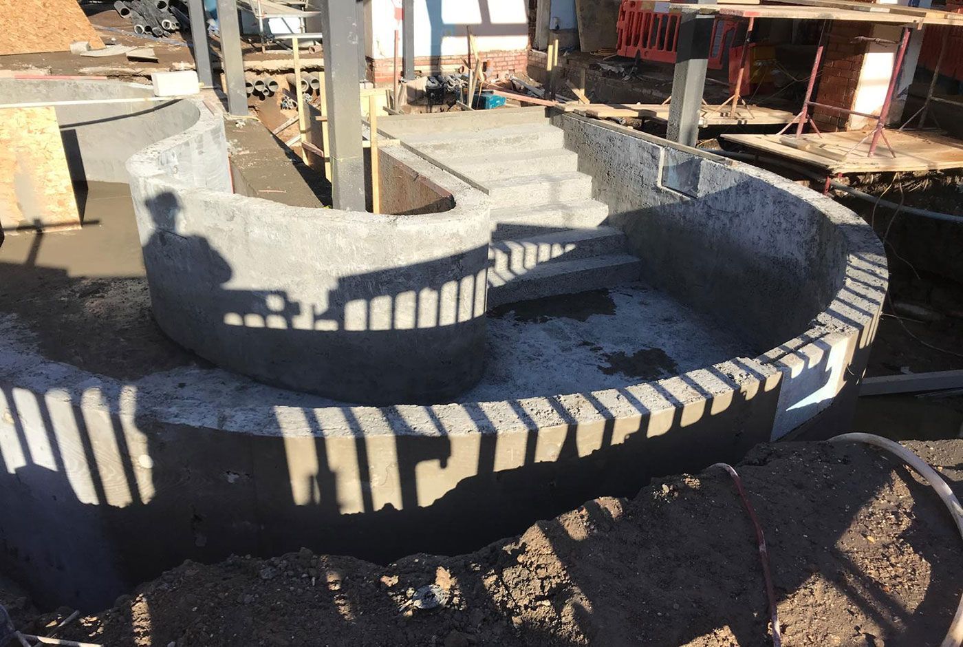 The concrete shell of an outdoor curved pool under construction, with steps down, in the shadow of a worker and railings