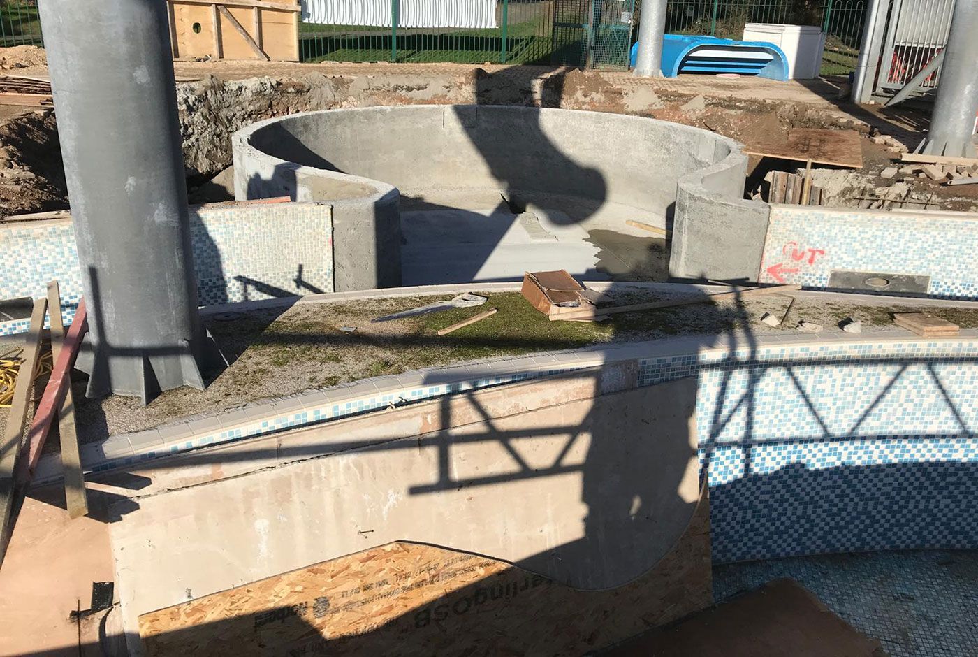 Concrete freeform pool shell under construction with unfinished walls, rebar, and a shadow across the structure.