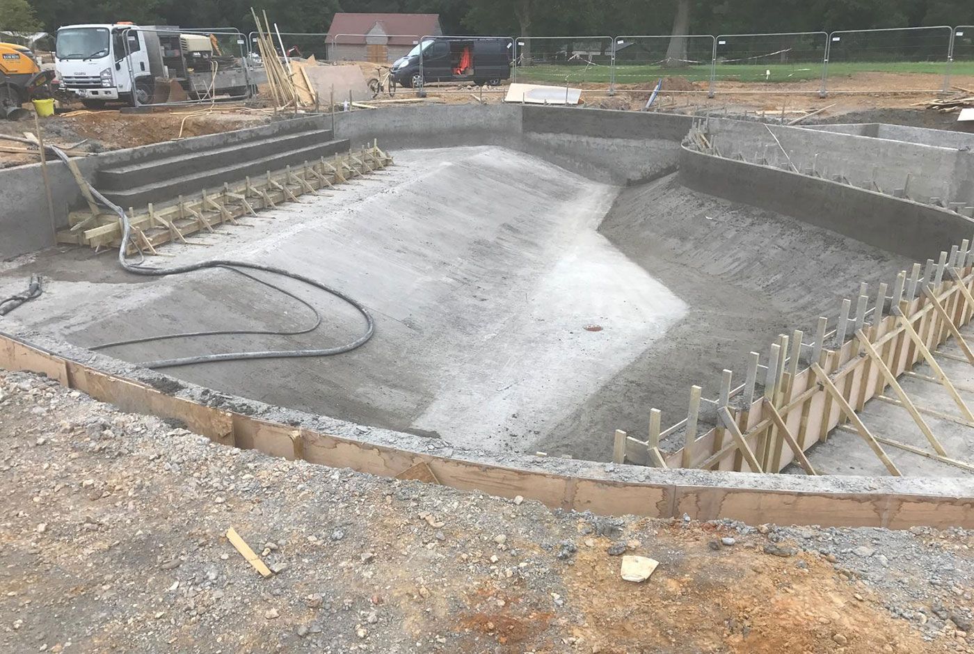 Outdoor swimming poo under construction with concrete walls, steps and sloping floor, fencing and vehicles in the background.