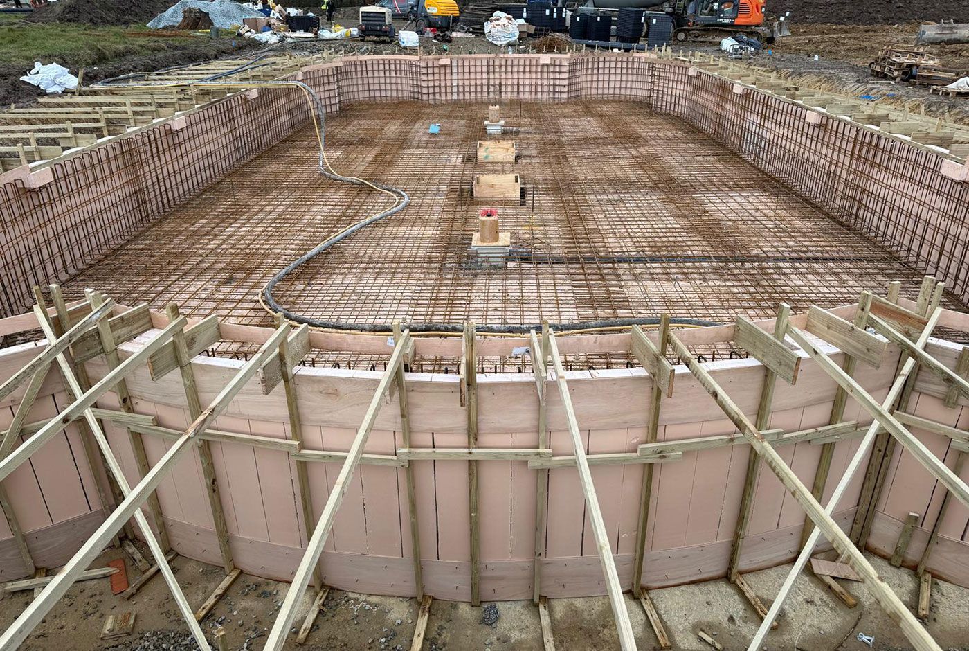 A large outdoor pool under construction, with rebar grid and wooden formwork, work equipment in the background.