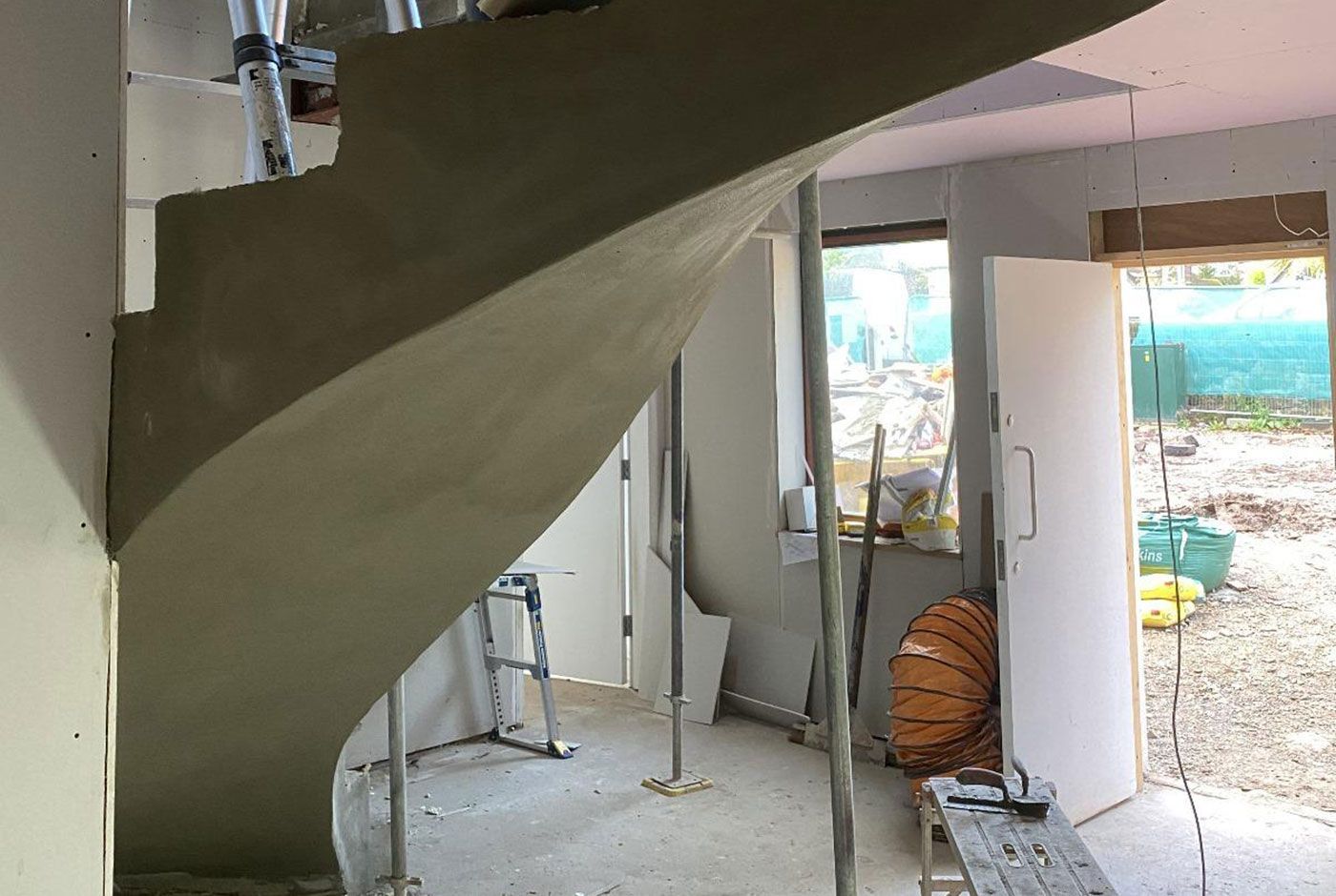 A concrete spiral staircase under construction within a building, doorway open to a bright outdoor area.