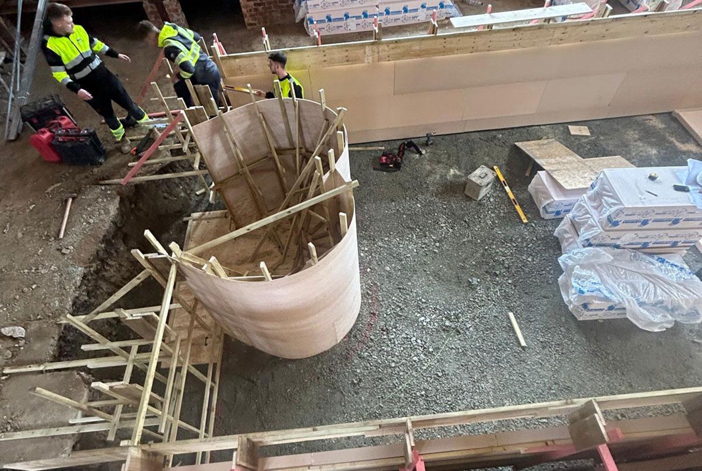 Workers constructing a large cylindrical form within the wooden frame of a swimming pool, with stacked materials nearby