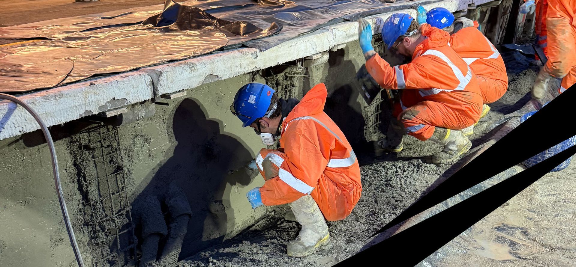 Workers in orange safety gear and blue helmets carrying out concrete repairs to a railway platform wall.