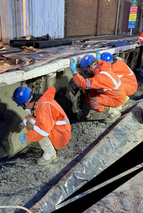 Workers in orange safety gear, blue helmets and gloves crouch inspecting a railway platform retaining wall that needs repair.