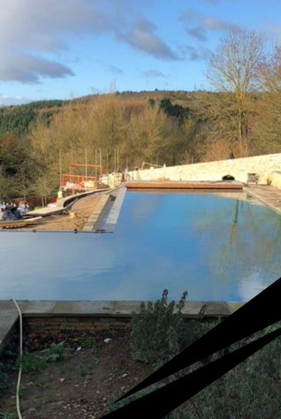 An outdoor infinity swimming pool under construction, with blue water, concrete walls, natural landscape in the background.