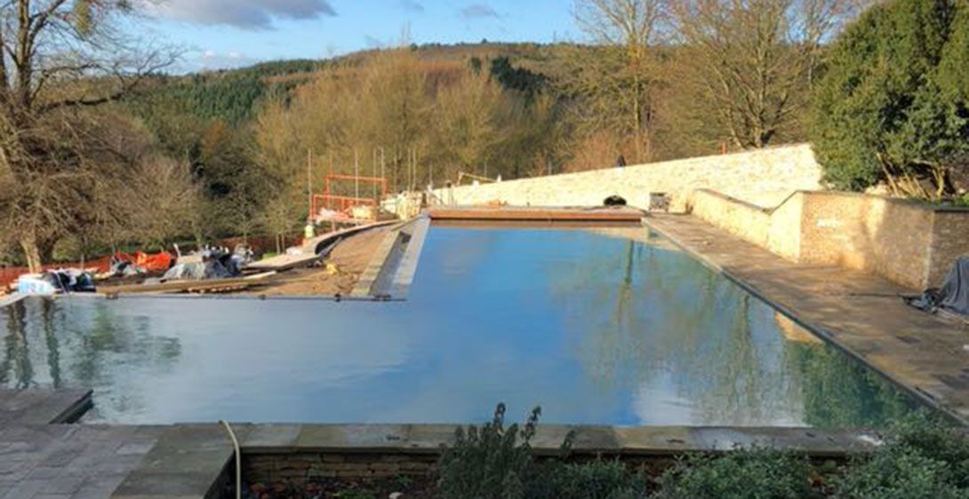 An outdoor infinity swimming pool under construction, with blue water, concrete walls, natural landscape in the background.