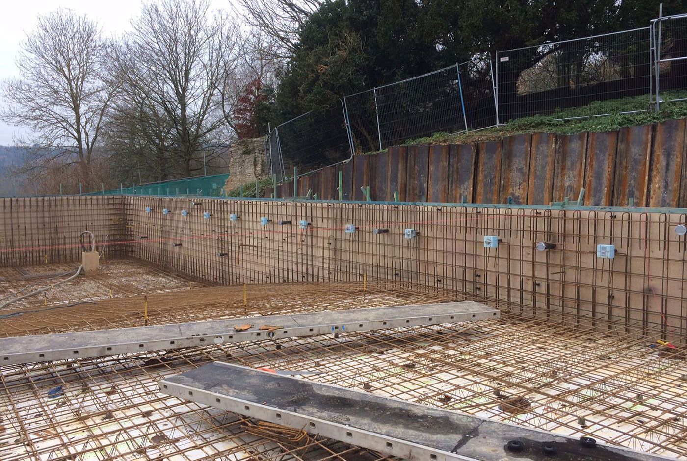 Excavated swimming pool onstruction site with timber retaining walls, rebar grid, and a steel-reinforced slope behind.