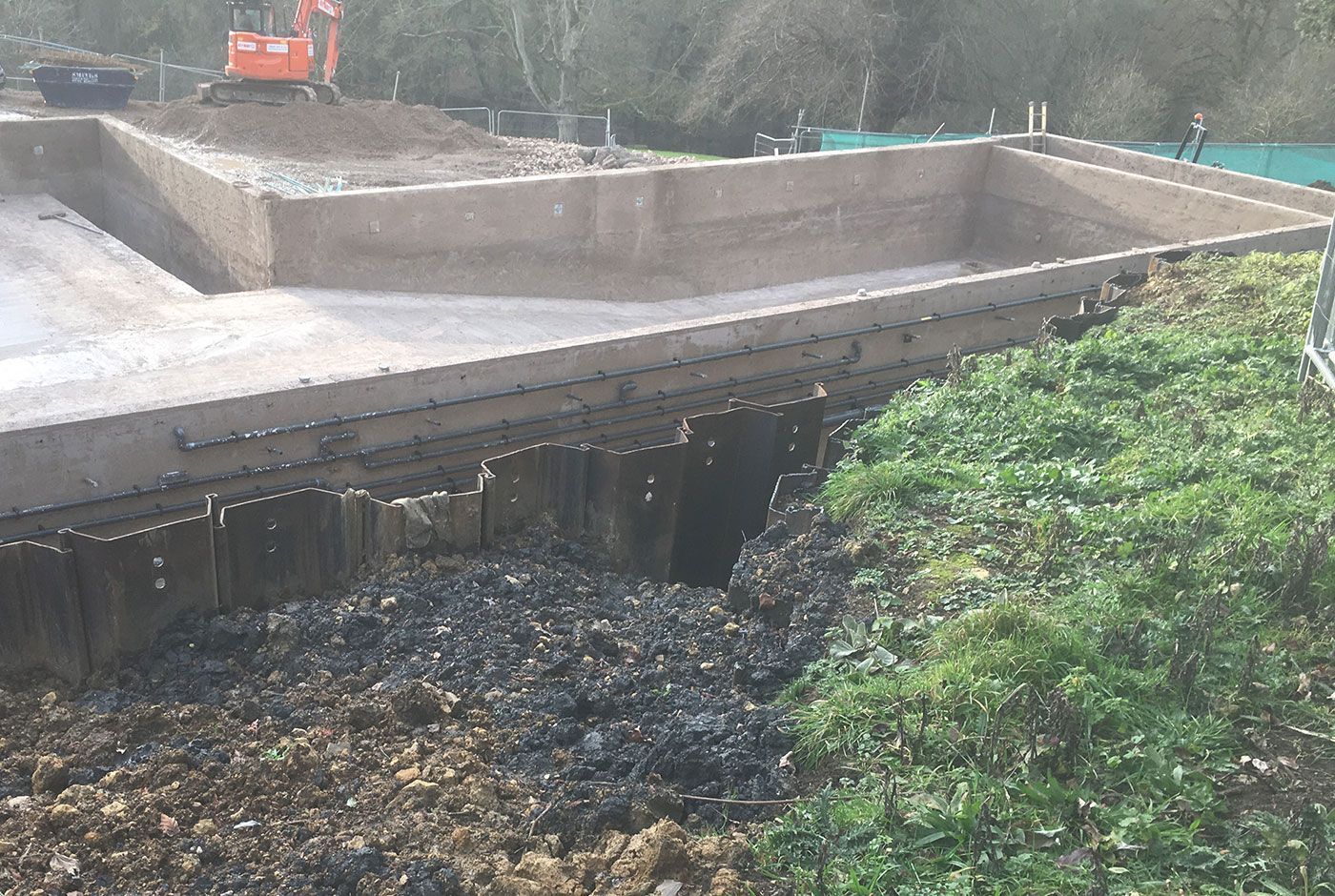 The concrete shell of a swimming pool with a mini excavator in the background and grass in the foreground.