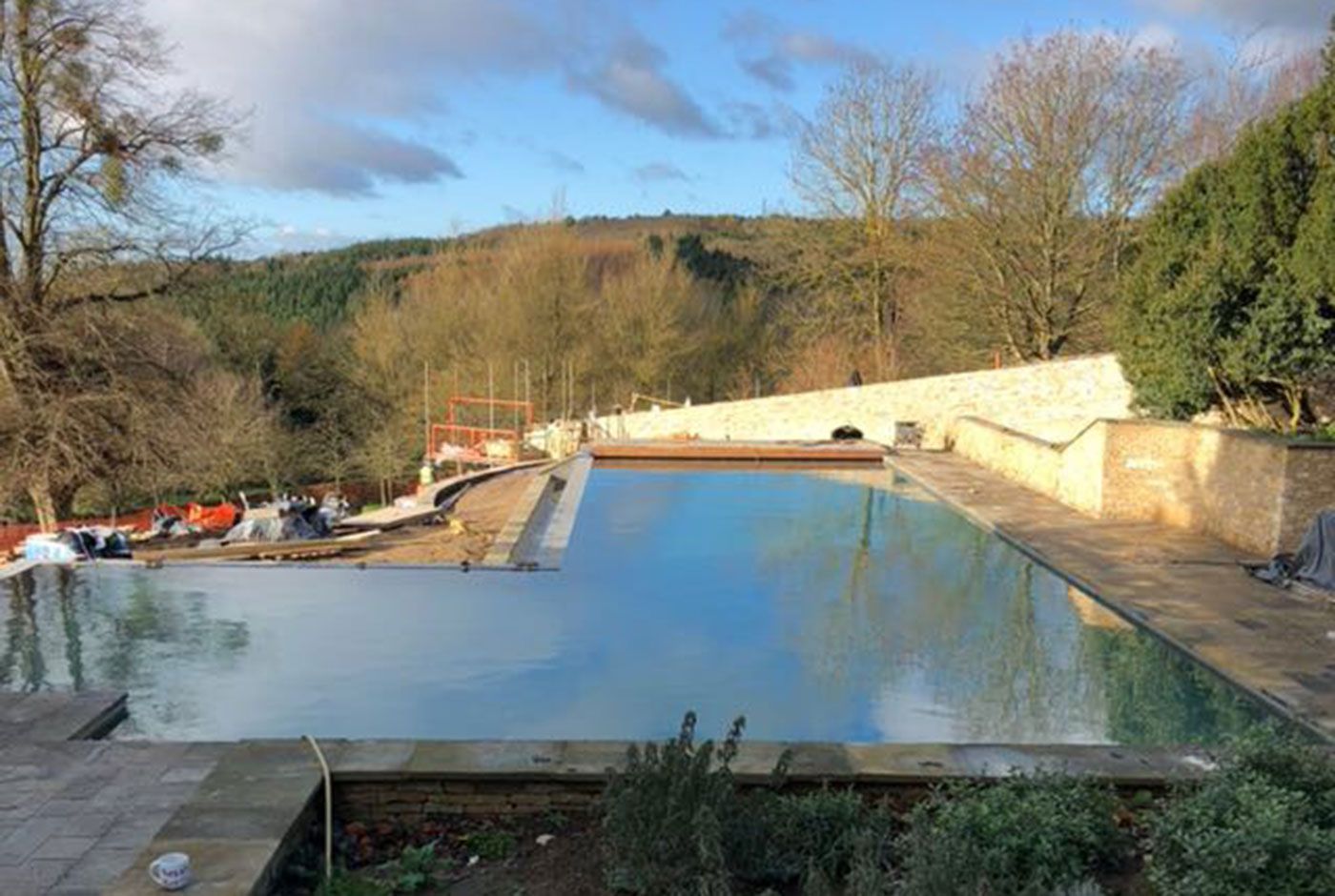 An outdoor infinity swimming pool under construction, with blue water, concrete walls, natural landscape in the background.