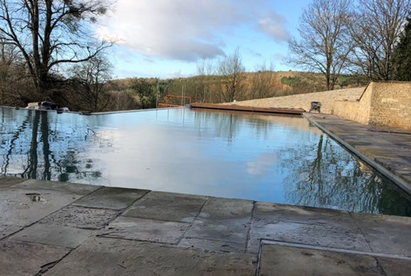 An infinity pool with a stone patio surround reflecting the blue sky with trees.