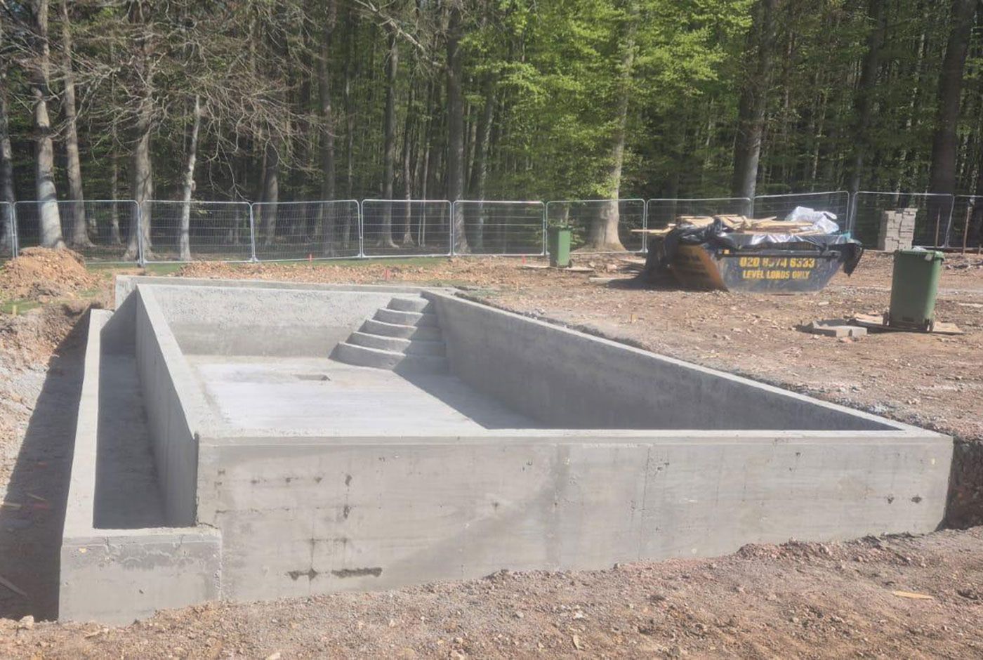 Concrete foundation for an infinity pool built on a wooded site, with fencing and construction equipment in the background.
